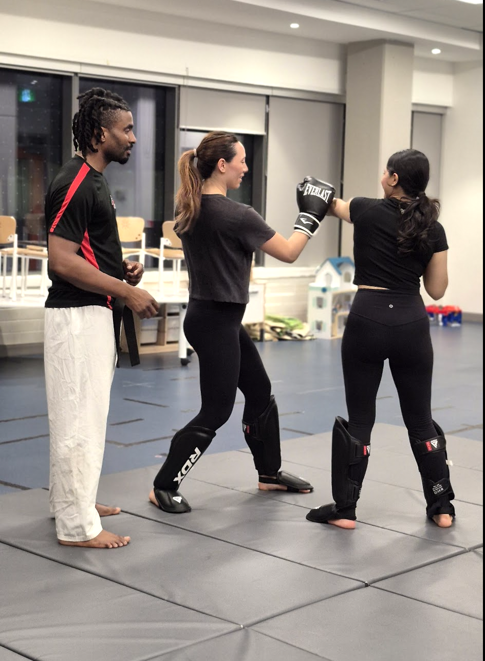 Women training self-defense with boxing gloves and shin guards under an instructor's guidance