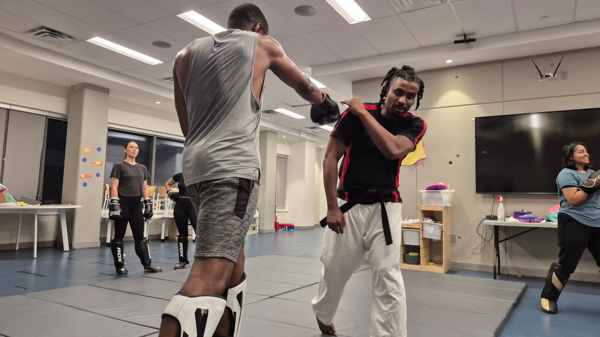 An instructor coaches an adult student through a padded sparring drill while classmates watch